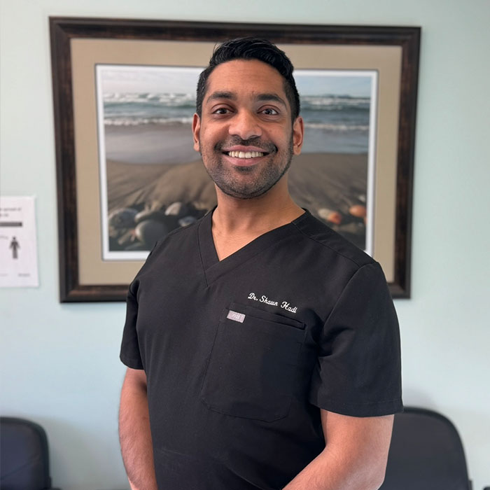 The image features a man in a medical scrubs uniform, standing indoors with a smile on his face. He is positioned in front of a framed picture on the wall.