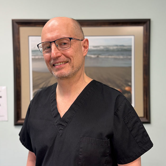 A bald man in glasses is smiling and standing next to a framed picture of a beach scene.