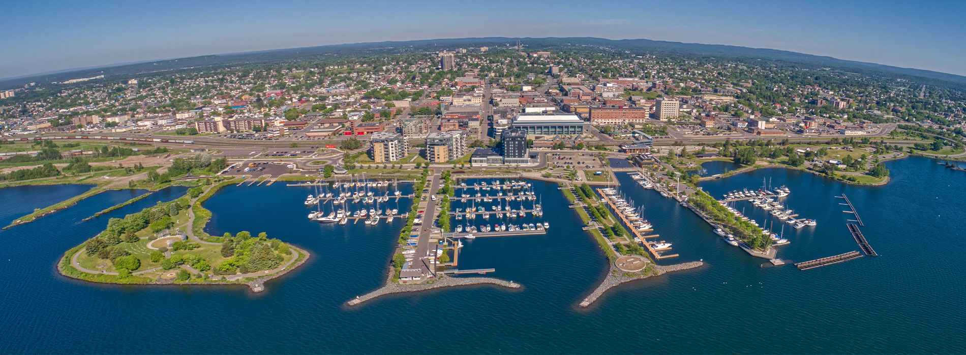 The image is a panoramic view of a harbor with multiple boats, a city skyline in the background, and a clear blue sky.