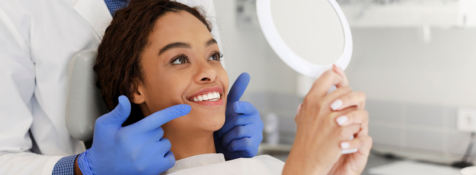 The image shows a woman seated in a dental chair, with her eyes closed and a smile on her face. She is holding a mirror and looking at her reflection. A dental professional is behind her, wearing gloves and a mask, indicating a dental care setting.