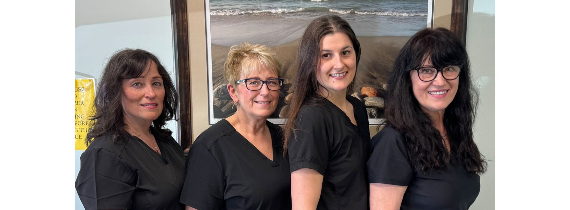 The image shows a group of five individuals, likely hairdressers or salon staff, posing for a portrait in front of a wall-mounted framed picture. They are dressed in professional attire and appear to be standing in an office or salon setting.