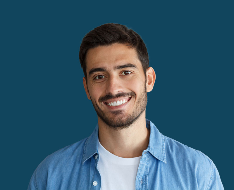 The image is a photograph of a young man smiling at the camera. He has short hair and appears to be in his mid-twenties. He is wearing a blue denim shirt and has a beard. Behind him, there's a plain background with a gradient from light to dark blue.