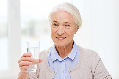 The image shows an elderly woman holding a glass of water to her mouth, smiling slightly.