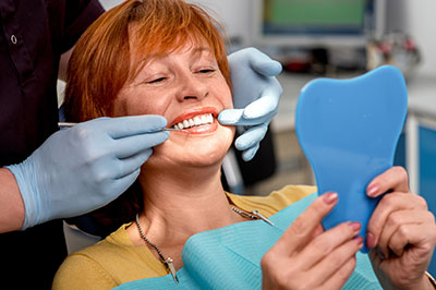 The image shows a woman in a dental chair, smiling at the camera, with her teeth being worked on by a dental professional.