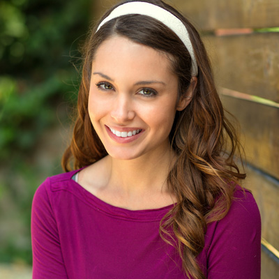 A woman with long brown hair, wearing a purple top and a headband, smiling at the camera.