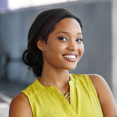 A young woman with a bright smile, wearing a yellow top and a necklace, posing against a blurred background.