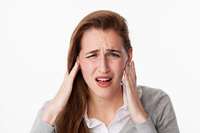 Woman in a white shirt and grey sweater holding her head with both hands, expressing concern or pain.