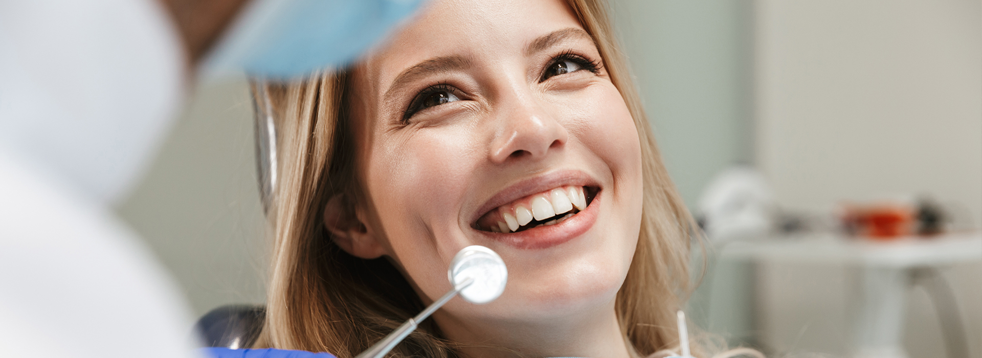 The image is a photograph of a young woman with a smile, sitting in front of a dental chair, being attended to by a dentist.