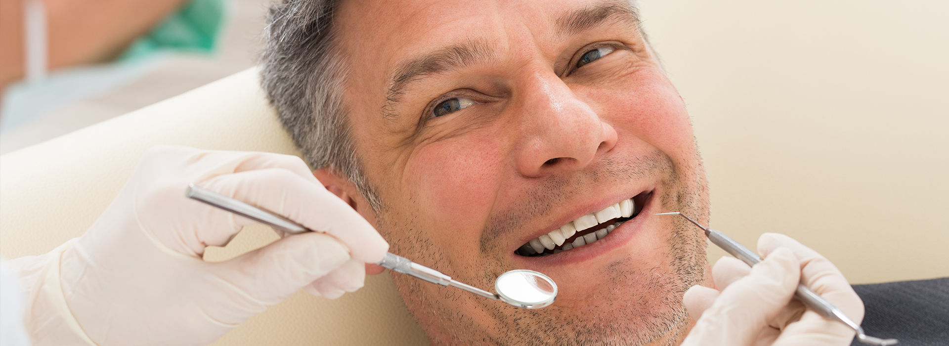 A smiling man in a dental chair receiving dental care, with a dentist holding a tool and wearing gloves.