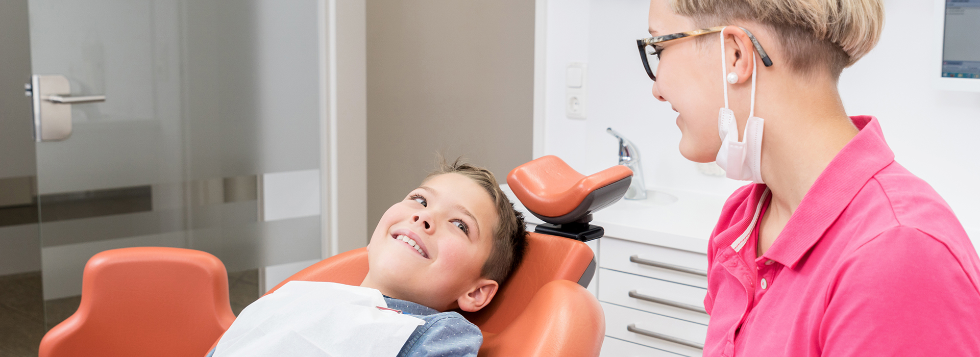 The image shows a dental office setting with a young boy in the chair, a dentist standing beside him, and another person observing from behind.