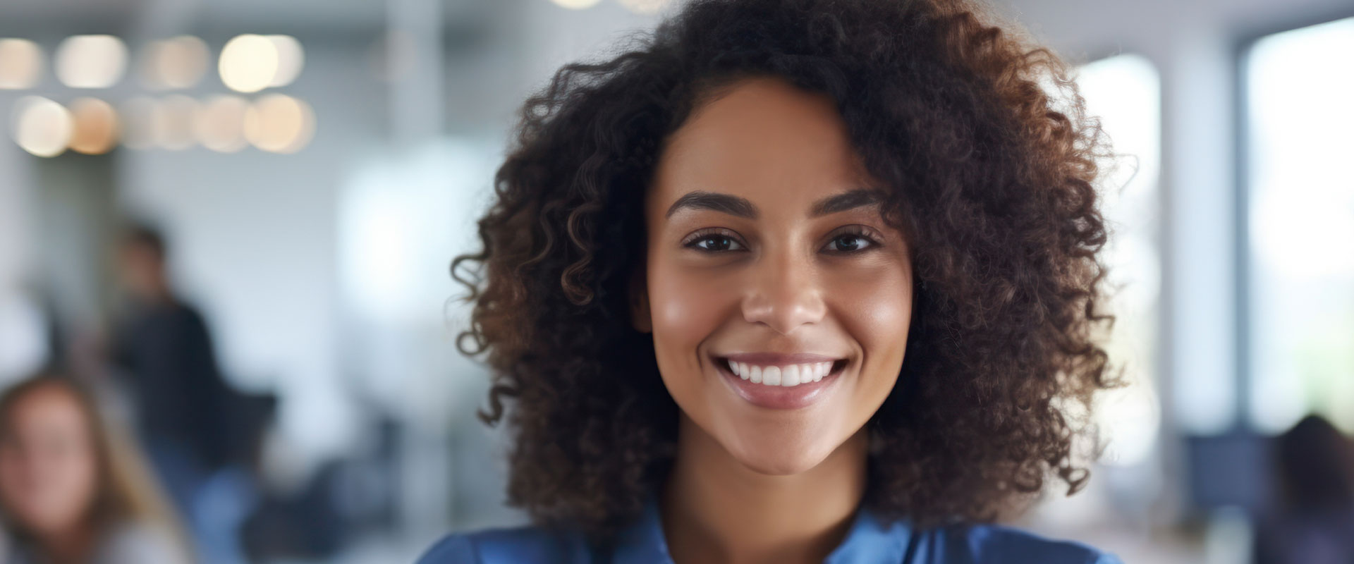 The image shows a person smiling at the camera, set against an office background with other individuals present.