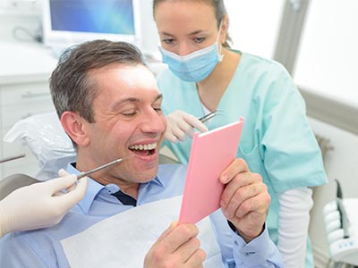 Man in a dental chair, smiling and showing a pink card with a surprised expression, while a dentist looks on.