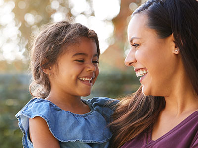 The image shows a woman and a young child, both smiling and looking towards the camera. They appear to be outdoors in a park or similar setting.