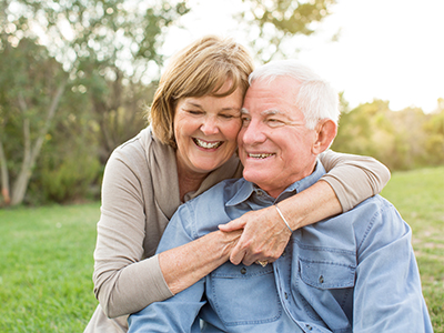 The image shows an elderly couple, a man and a woman, embracing each other in a warm, affectionate manner. They are outdoors during what appears to be either dawn or dusk, given the soft lighting, and they are both smiling at the camera.