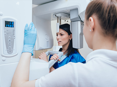 Image  A woman in a blue lab coat stands next to a large, modern MRI machine with digital displays, while another person observes from behind.