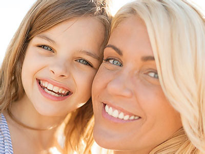 The image shows a woman with blonde hair smiling at the camera, holding a young girl with brown hair who is also smiling. They appear to be outdoors during daylight hours.