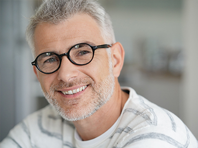 The image is a photograph of a middle-aged man with grey hair, wearing glasses and a white shirt. He has a beard and mustache, and he is smiling at the camera.