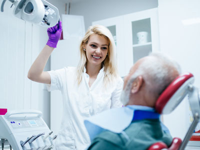 A dental professional in a white coat and gloves is standing next to a patient in a hospital bed, who appears to be receiving dental care.