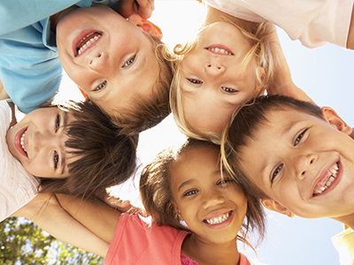 The image shows a group of children of varying ages, with some smiling and looking directly at the camera, while others are slightly turned away or looking upwards. They appear to be outdoors on a sunny day, as indicated by the bright lighting and shadows.