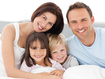 A family of four, including two adults and two children, in a bedroom setting with a smiling father, mother, and two children.