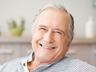 The image shows a smiling older man with gray hair, wearing a blue shirt and seated in a relaxed posture.