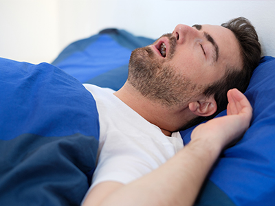 The image shows a man lying in bed with his eyes closed, appearing to be asleep or resting with his head on a pillow.