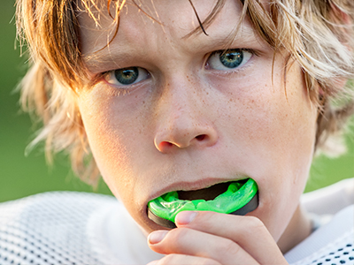 The image depicts a young male with blonde hair and blue eyes, wearing a football jersey. He is holding a green object in his mouth that resembles a toothbrush or a similar dental hygiene tool.