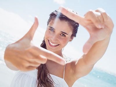 A woman is taking a selfie with her left hand, smiling, and holding up her index finger in front of the camera. She is outdoors, under clear skies, with a sunny backdrop featuring the ocean.