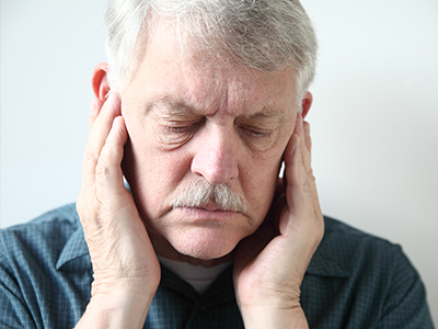 The image shows an older man with a mustache, wearing glasses and a blue shirt, holding his hand to his ear in a gesture of pain or discomfort.